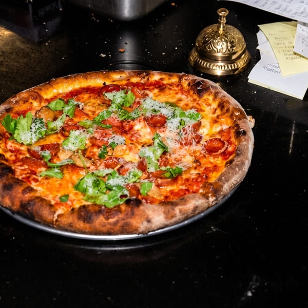 A freshly baked pizza with pepperoni, cheese, and green herbs sits on a dark countertop next to a golden bell.