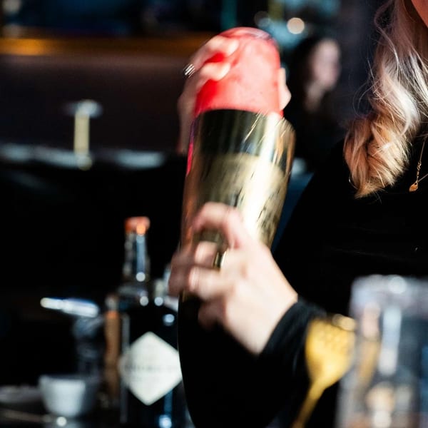 Bartender shaking a cocktail in a gold shaker, with blurred bar lights in the background.