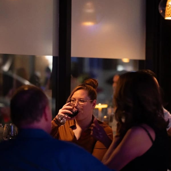 A woman in glasses sips red wine at a dimly lit restaurant table with other patrons.