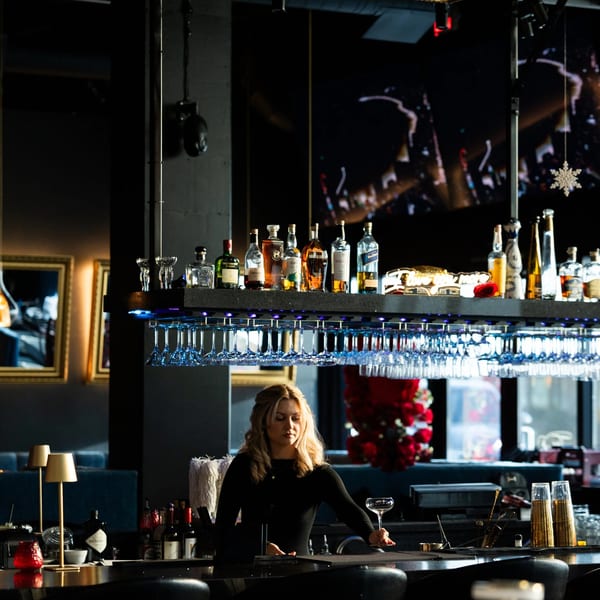 Blonde bartender in a black shirt holding a coupe glass behind a well-stocked bar.