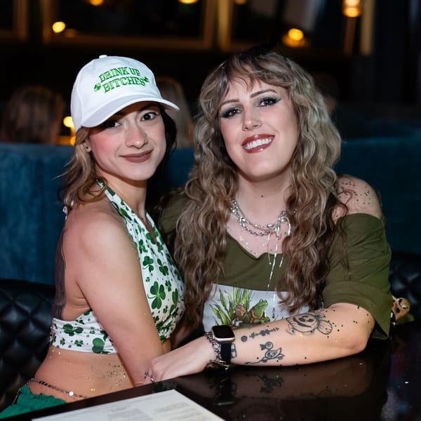 Two women in festive green attire with glitter, smiling at a table in a dimly lit restaurant.