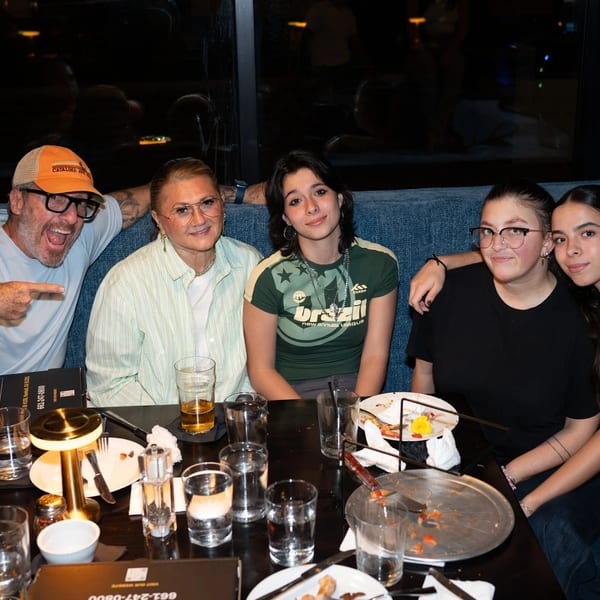 Five people, including a man pointing, seated at a restaurant table with drinks and pizza.