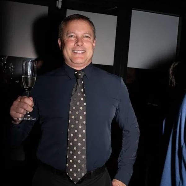 A smiling man in a dark shirt and polka-dot tie holds a glass of champagne.