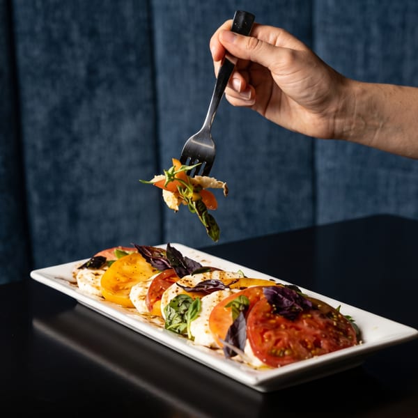 Hand holding a fork with food over a caprese salad with red and yellow tomatoes.