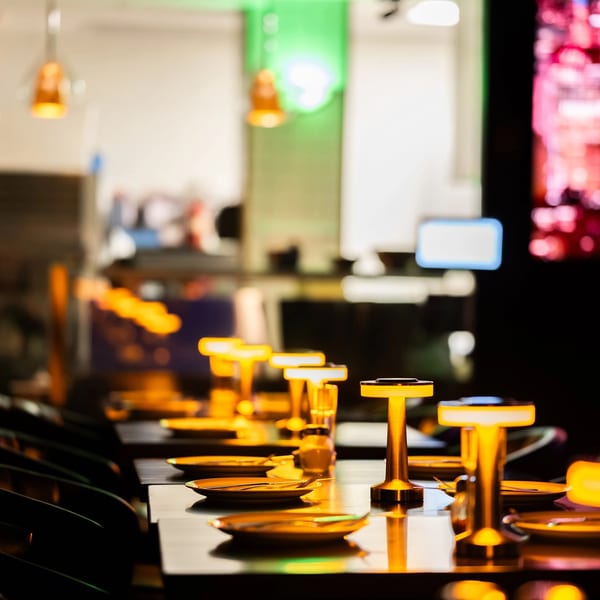 A row of restaurant tables set with plates, cutlery, and glowing gold lamps in a dimly lit room.