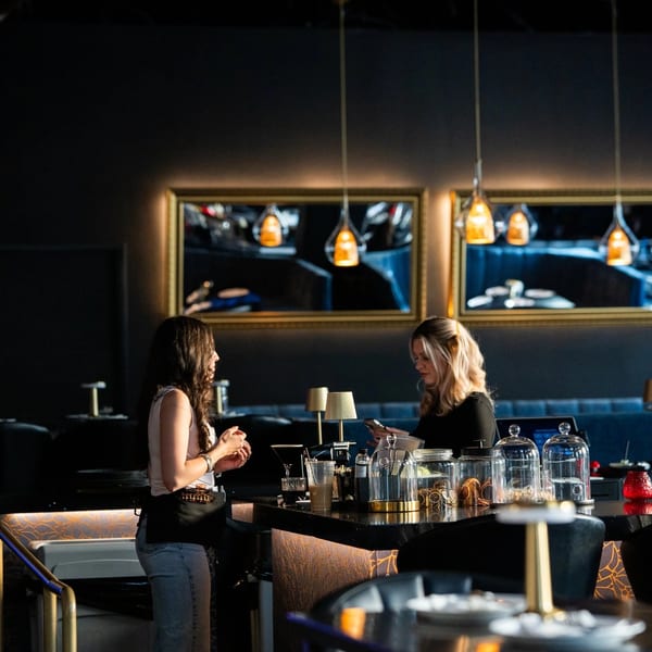 Two women conversing at a dark, modern bar with gold accents and pendant lights.