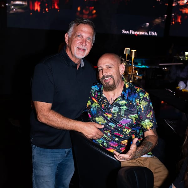 Two men smiling at the camera, one in a black polo, the other in a colorful mushroom shirt.