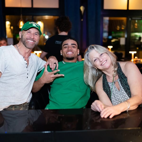 Three smiling people, two men and one woman, pose together at a dark restaurant bar.