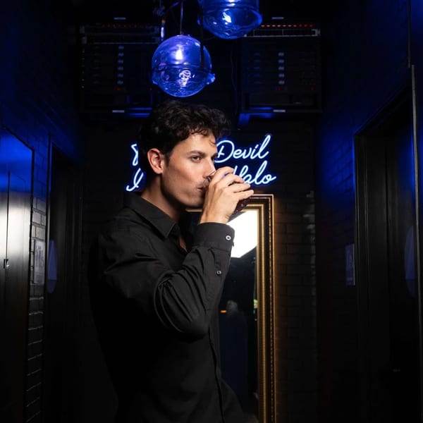 Man in a black shirt sips a drink in a dark, blue-lit bar with a neon sign.