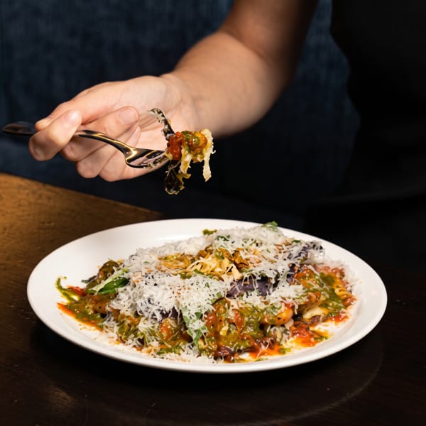 A hand holding a fork with a bite of pasta, covered in sauce and cheese, above a white plate.