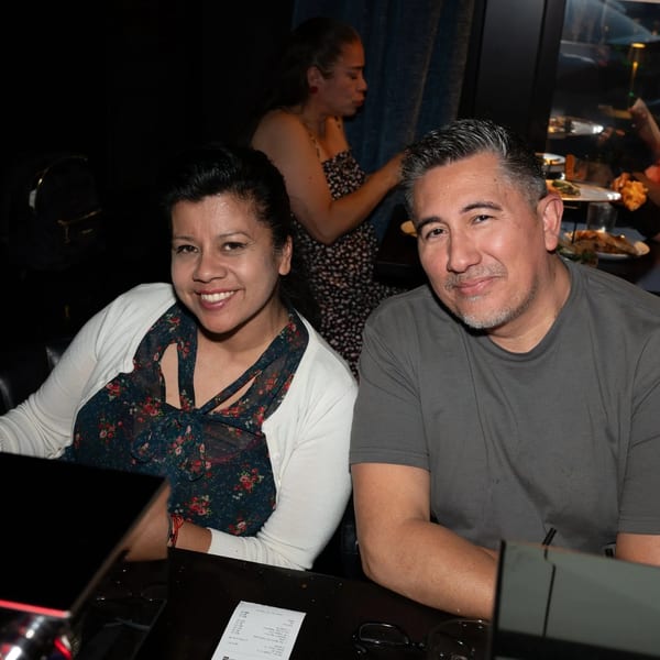 Two smiling people, a woman and a man, seated at a dark bar or table in a restaurant.